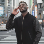 Ein stilvoller Mann blickt in einer belebten Großstadt lächelnd nach oben, während er die schwarze SQUEQO Urban Hybrid Bomberjacke mit Lederärmeln trägt.