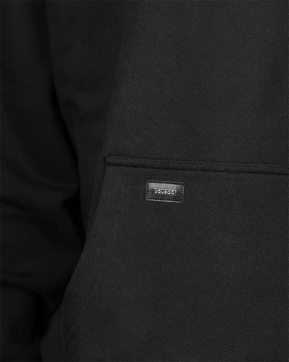 Detailaufnahme des hochwertigen Metall-Logo-Patches mit SQUEQO-Schriftzug, platziert auf der Kängurutasche des schwarzen Oversized Hoodies.