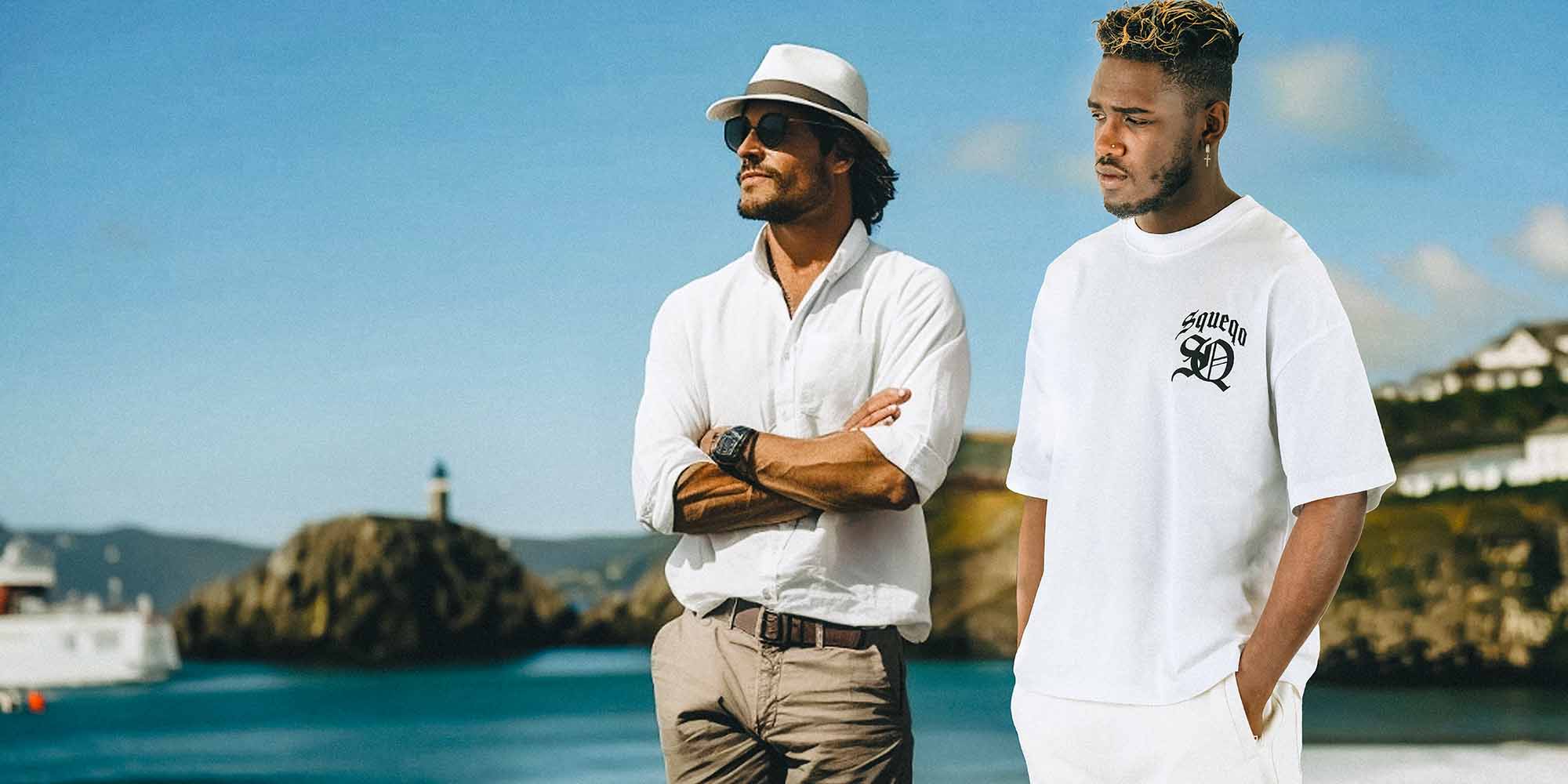 Zwei Männer in Sommeroutfits stehen an einem Küstenort, einer in einem weißen Hemd, Hut und Sonnenbrille, der andere in einem weißen T-Shirt mit Squeqo-Monogramm, beide blicken in die Ferne.