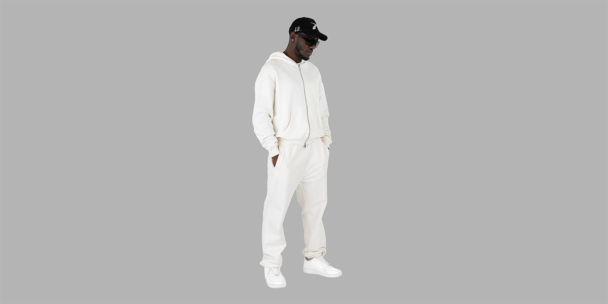 Mann in stilvollem weißen Zip-Hoodie und passender Jogginghose mit weißen Sneakern und schwarzer Cap steht seitlich auf grauem Hintergrund