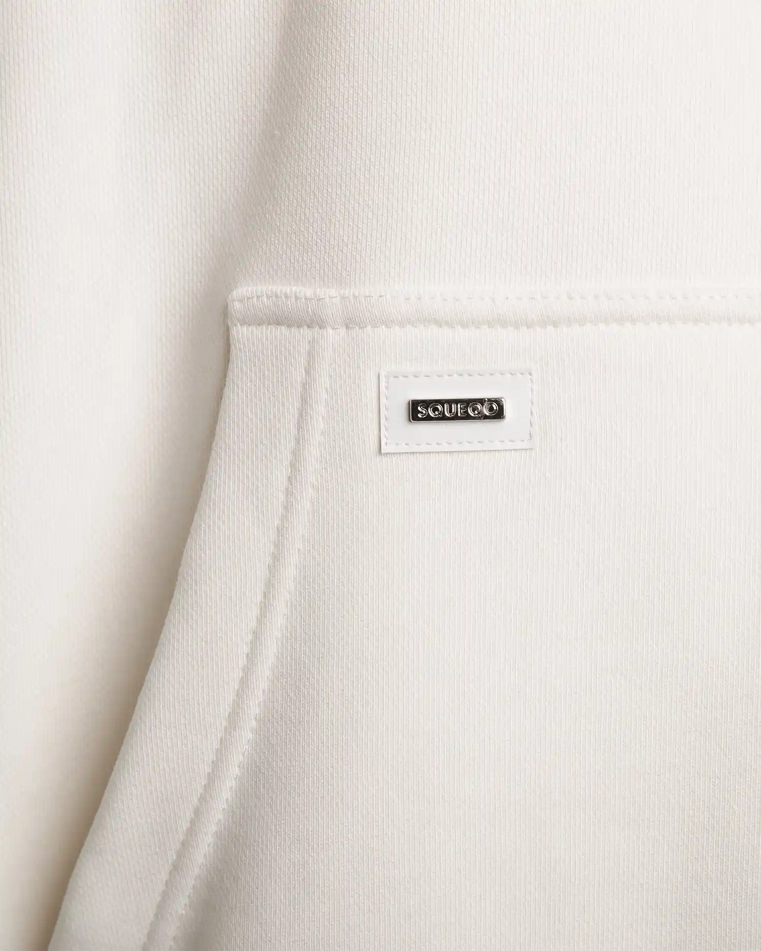 Detailaufnahme des Squeqo Metall-Patches an der Kängurutasche des Hoodies.