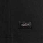 Detailaufnahme des Squeqo Metall-Patches mit Logo am Saum des schwarzen T-Shirts.
