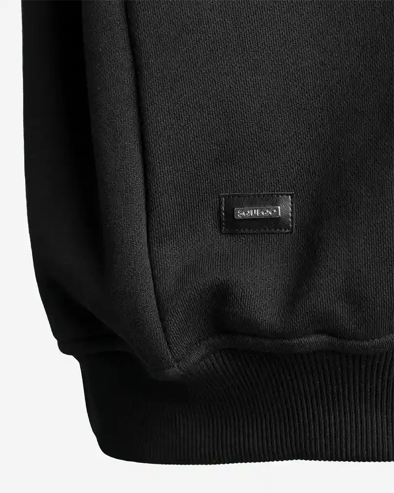 Detailansicht: SQUEQO Metall-Logo-Patch am Saum des Sweatshirts