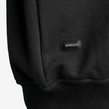 Detailansicht: SQUEQO Metall-Logo-Patch am Saum des Sweatshirts