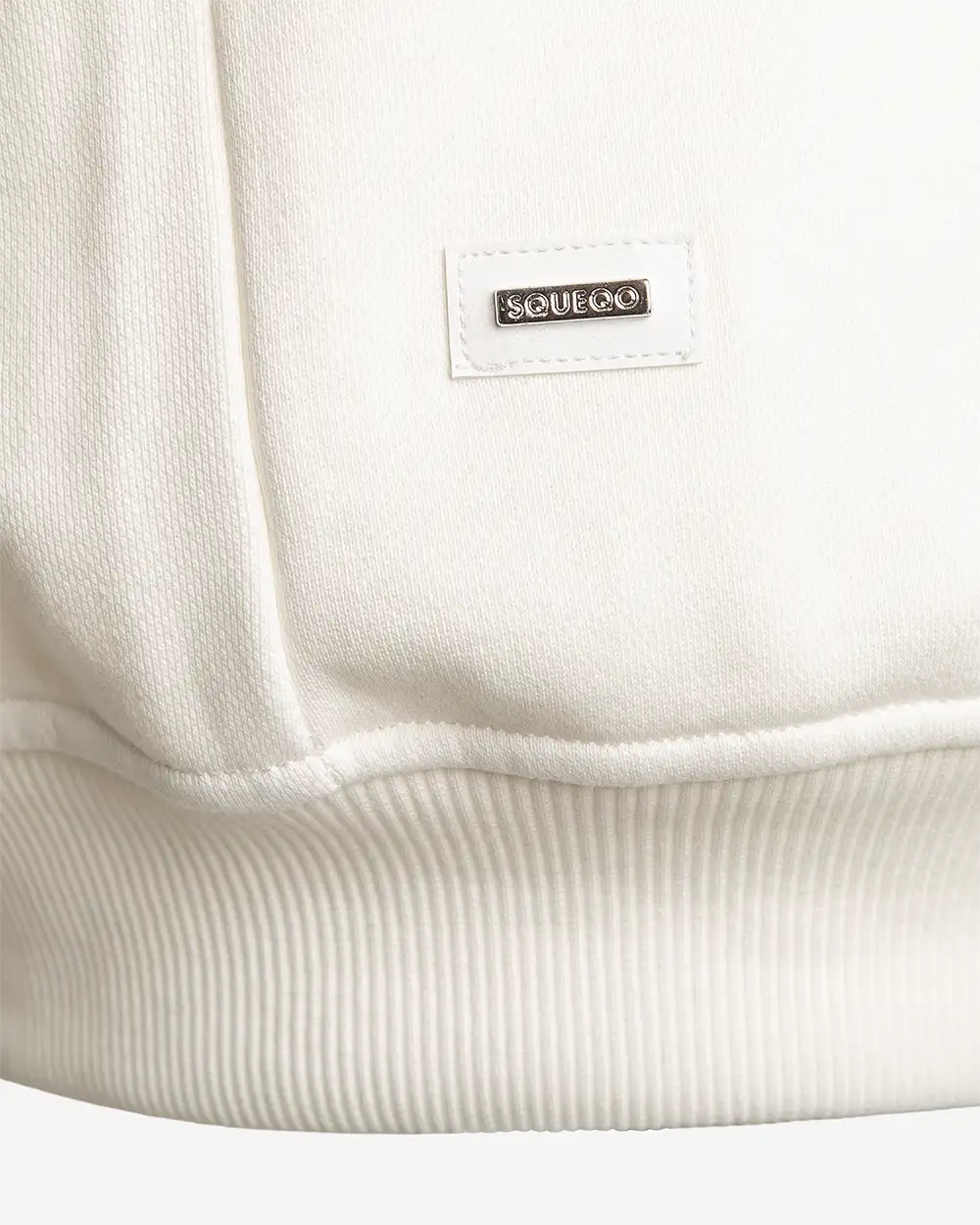 Detailansicht: SQUEQO Metall-Logo-Patch am Saum des weißen Sweatshirts
