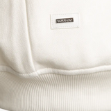 Detailansicht: SQUEQO Metall-Logo-Patch am Saum des weißen Sweatshirts