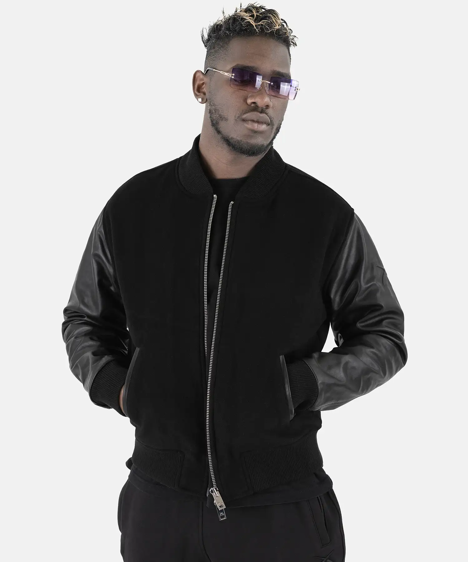 Studioporträt eines Mannes, der die schwarze SQUEQO Bomberjacke mit Lederärmeln trägt und mit Sonnenbrille selbstbewusst in die Kamera blickt.