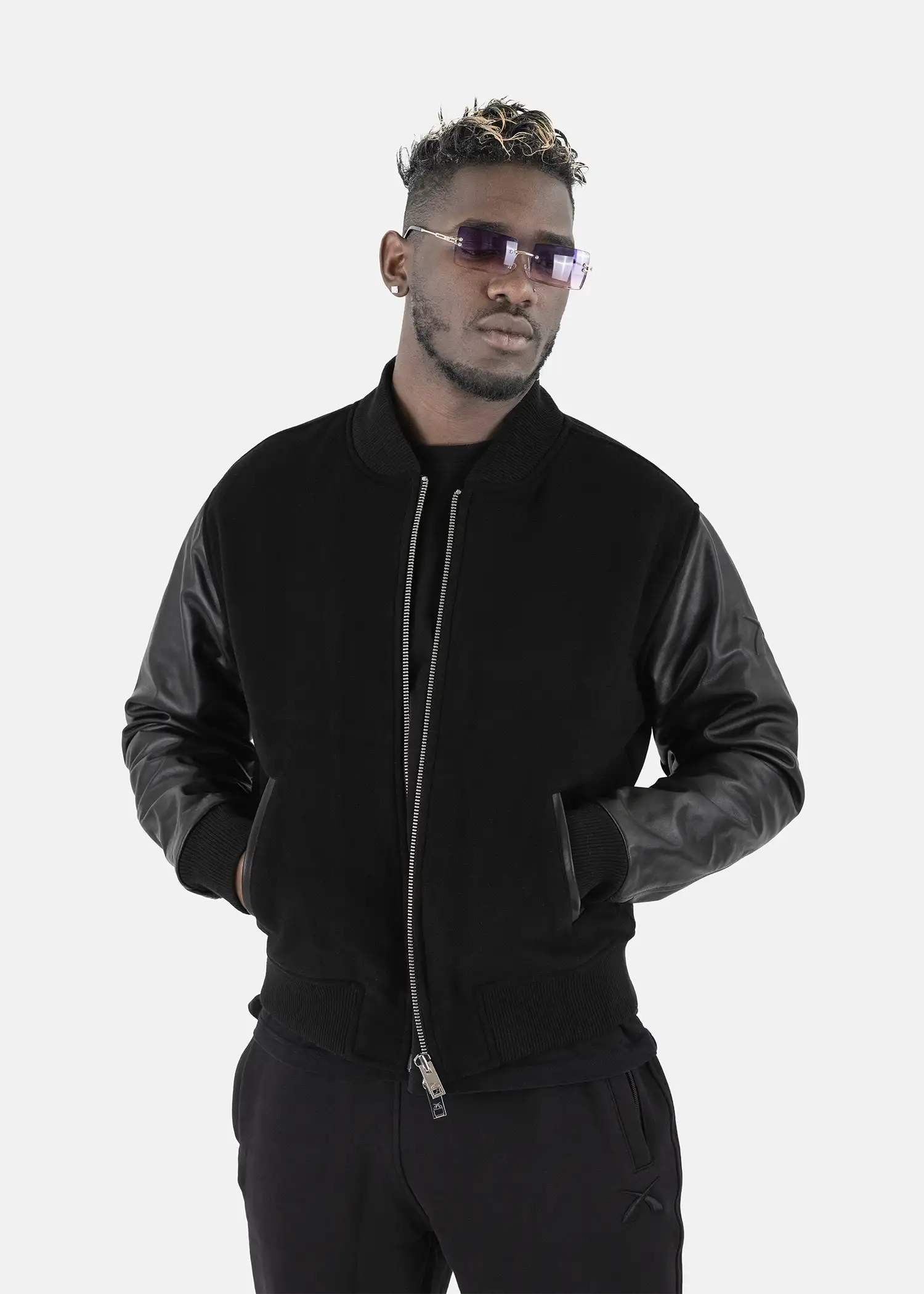 Studioporträt eines Mannes, der die schwarze SQUEQO Bomberjacke mit Lederärmeln trägt und mit Sonnenbrille selbstbewusst in die Kamera blickt.